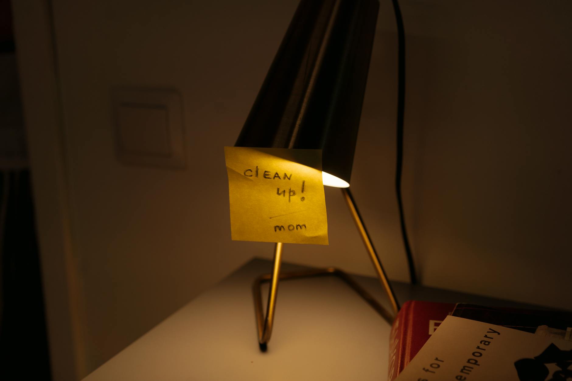 A desk lamp illuminates a sticky note with a \'clean up\' reminder in warm, cozy lighting.