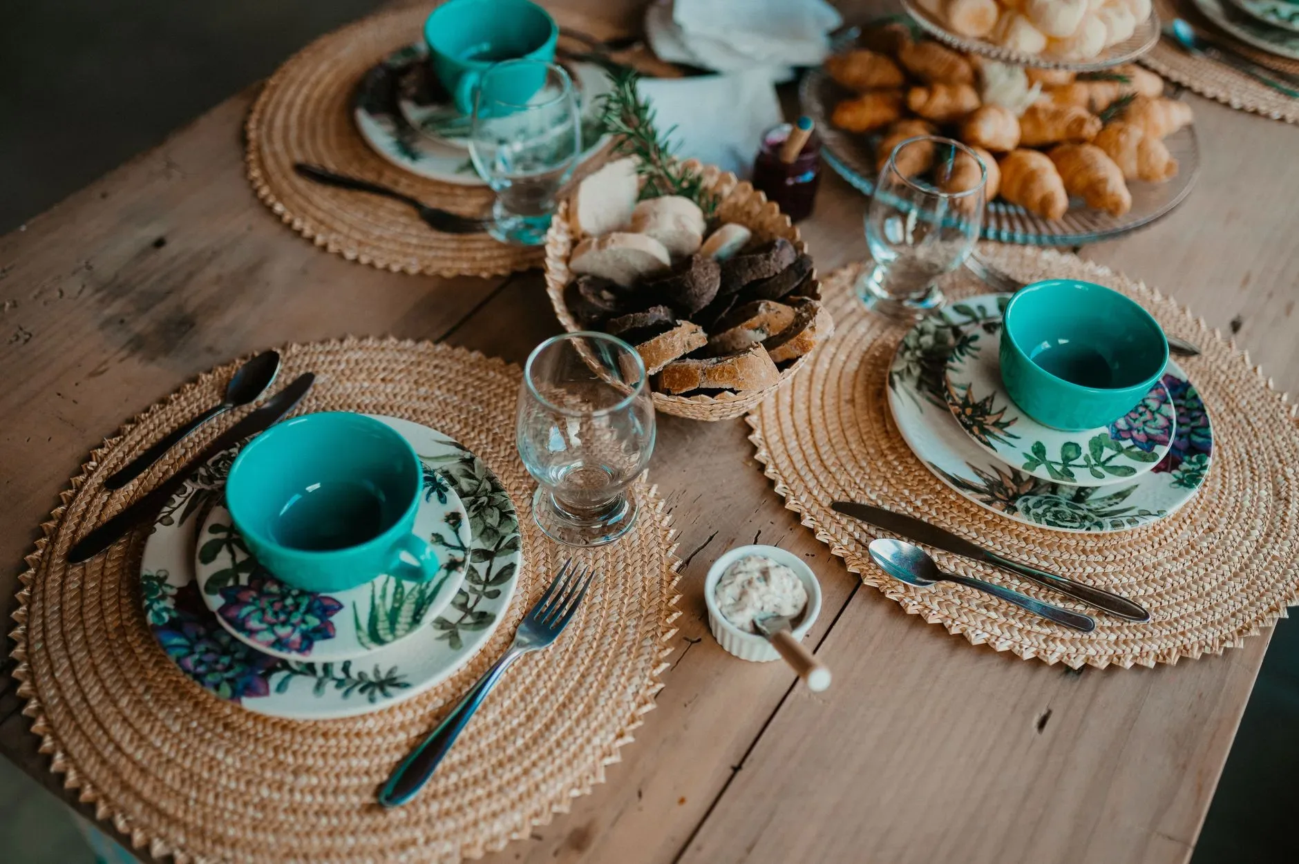 A rustic dining table set for a meal with bread, pastries, and floral plates creating a cozy atmosphere.