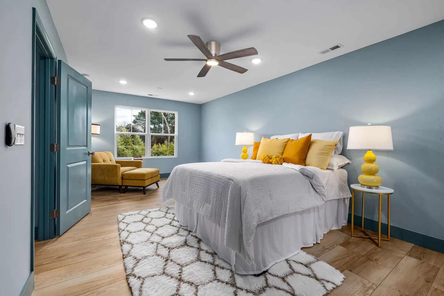 A stylish bedroom with yellow accents, featuring a bed, armchair, and ceiling fan.