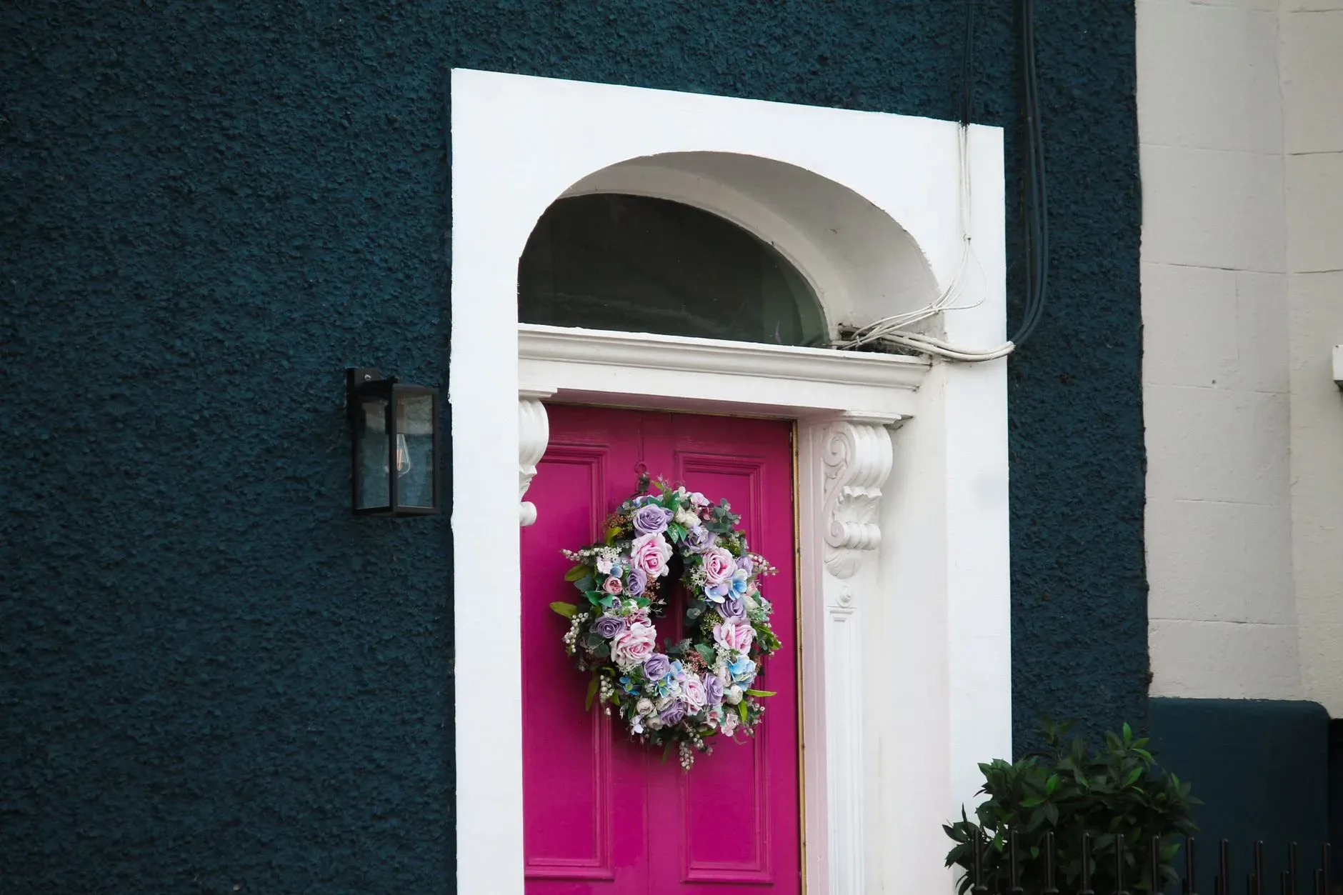 A vibrant pink door adorned with a floral wreath, set against a textured blue wall, exudes charm and elegance.