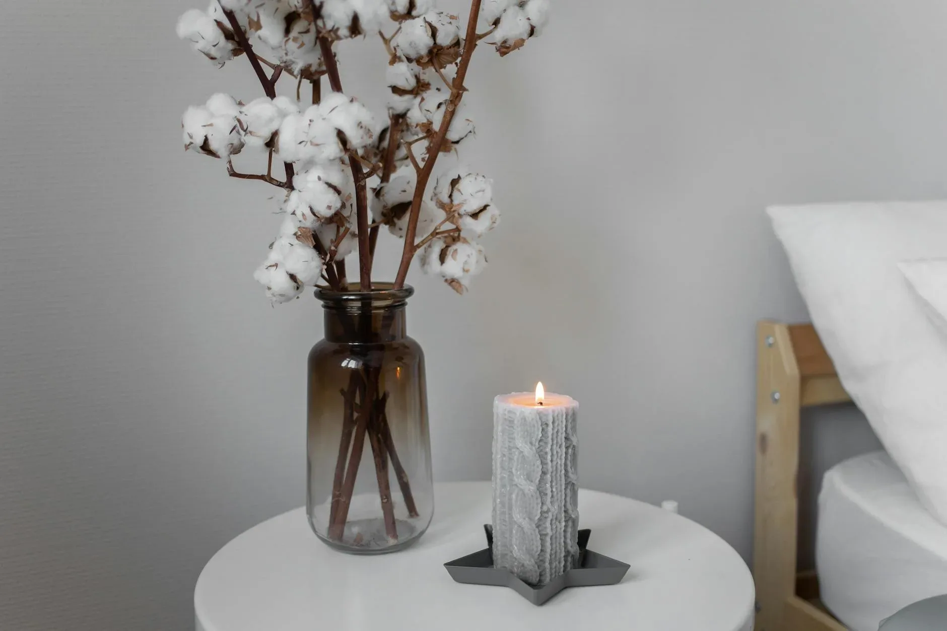 A warm and inviting still life with cotton stems in a glass vase and a lit candle on a bedside table.