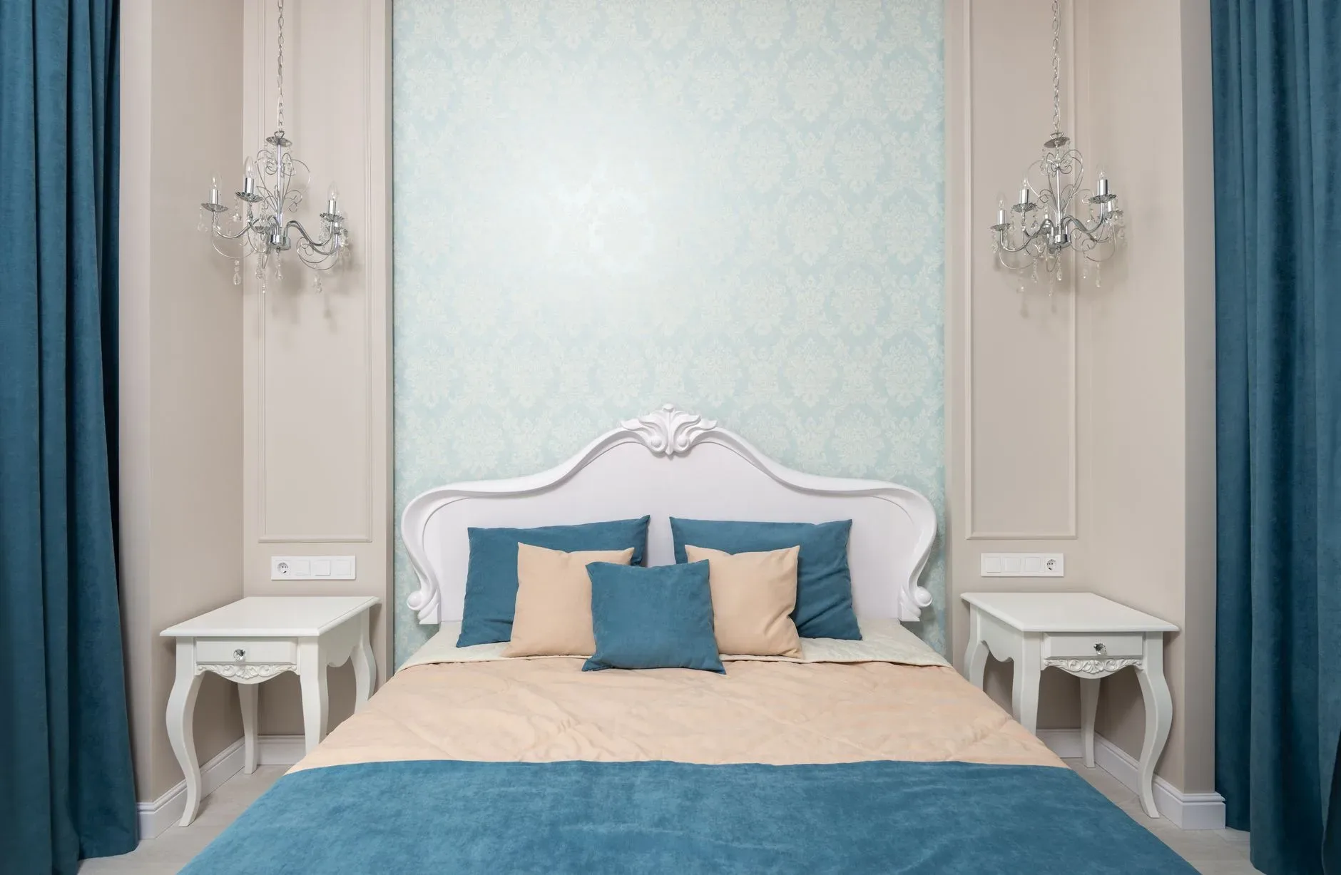 Comfortable bed with blue and beige bedclothes between bedside tables at wall with wallpaper and hanging lamps in bedroom with classic design
