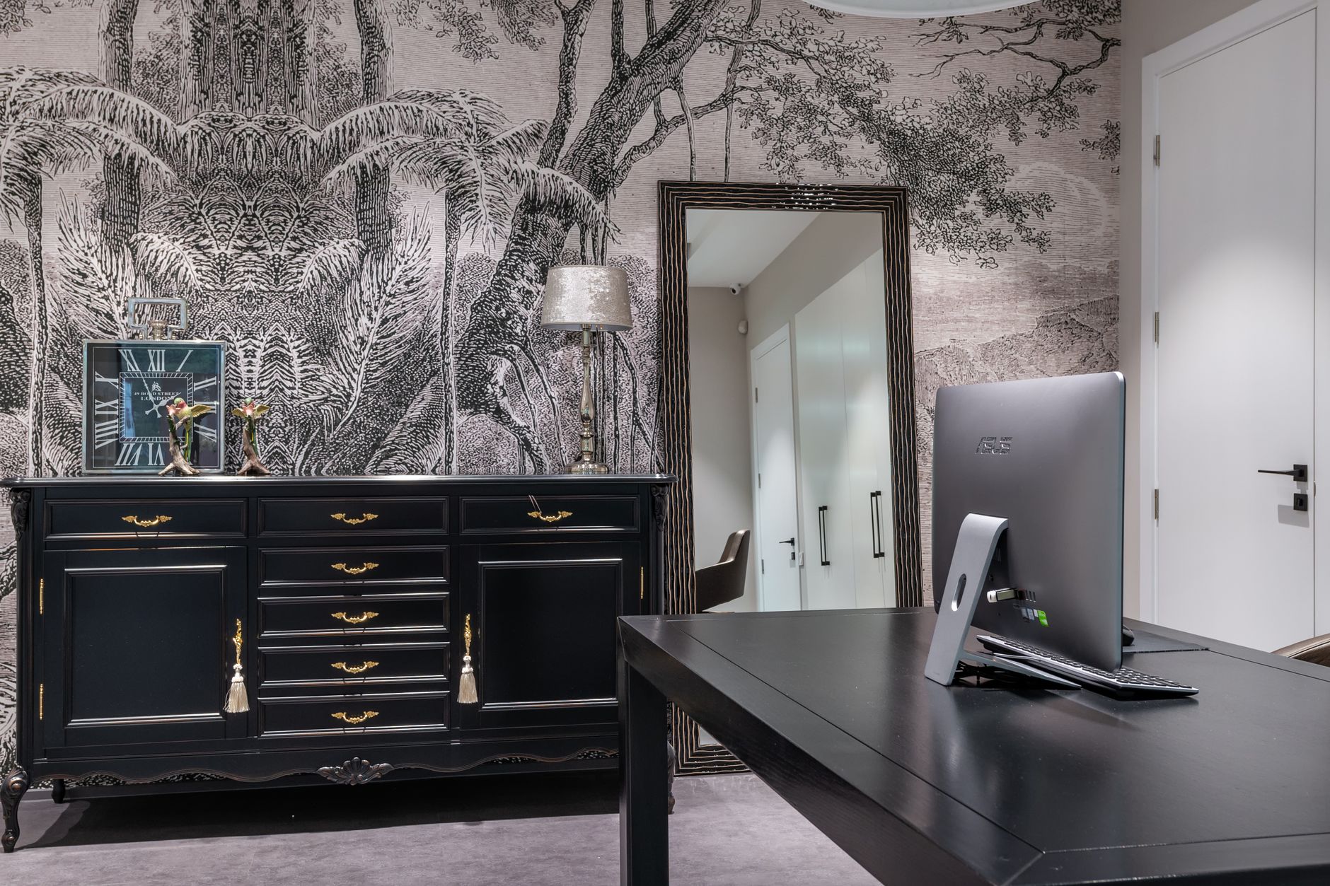 Contemporary computer on black table placed near cabinet with clock at wall with wallpaper and mirror in apartment with door