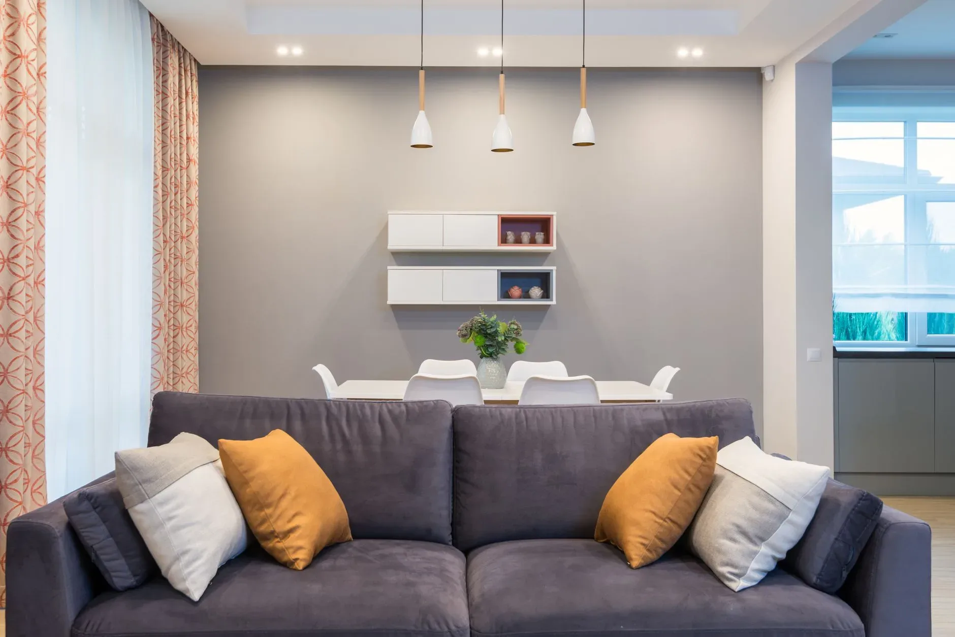 Soft sofa with cushions placed near table with chairs at wall with decorations on shelves in spacious flat with lamps