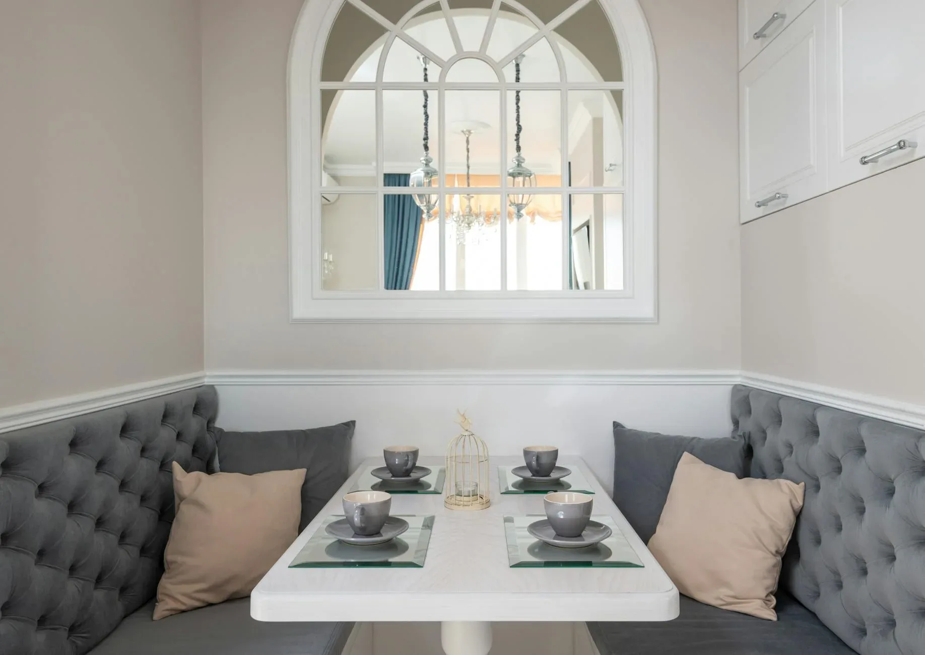 Table setting with mugs and plates near soft settees with pillow at wall with window in modern dining room of apartment