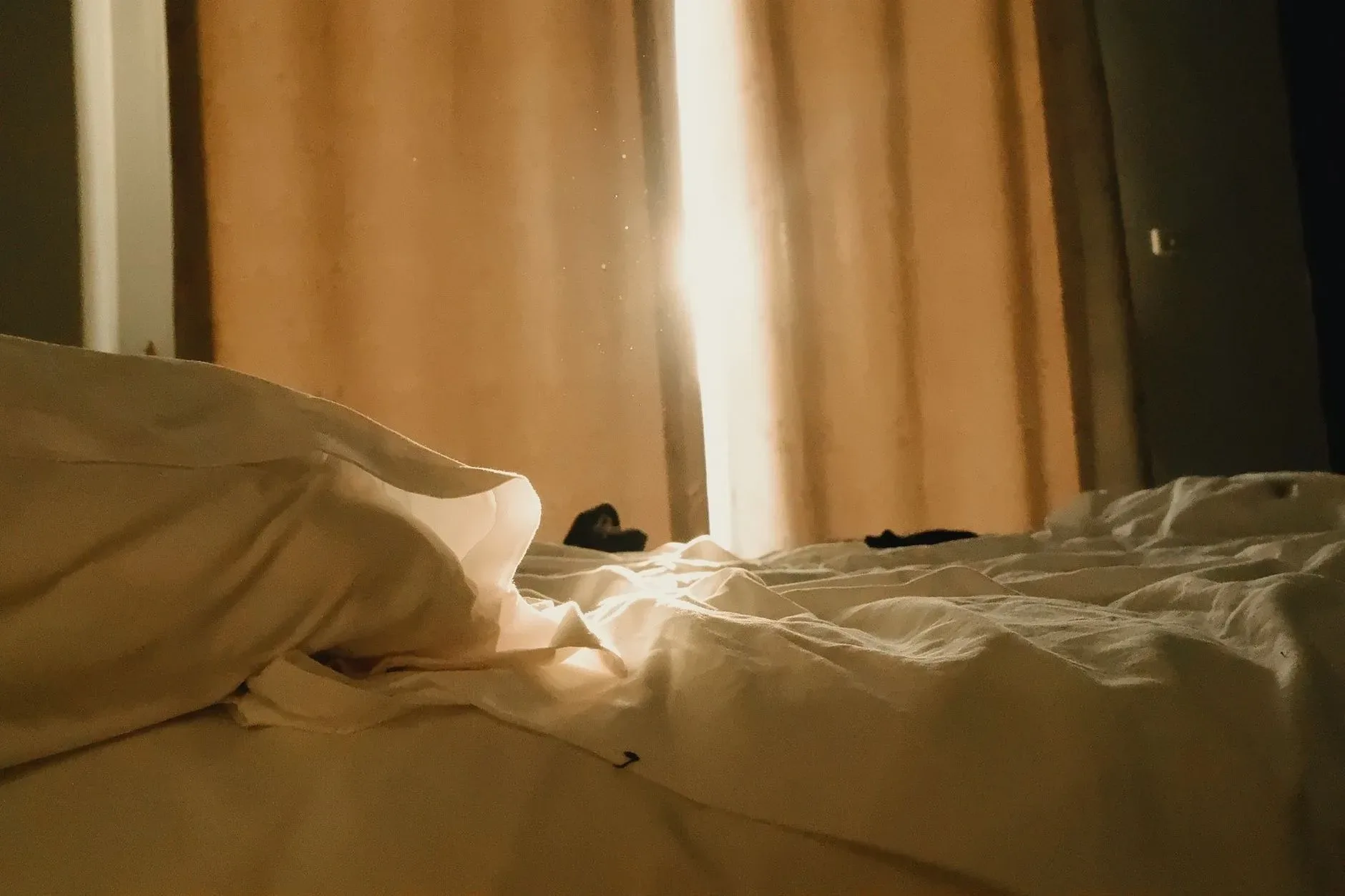 Warm sunlight streaming through curtains onto a cozy, unmade bed in a tranquil room.